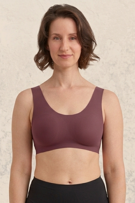 A woman modeling a sleeveless scoop-neck bra in a dusty mauve-brown color, paired with black athletic bottoms, set against a textured light beige background.