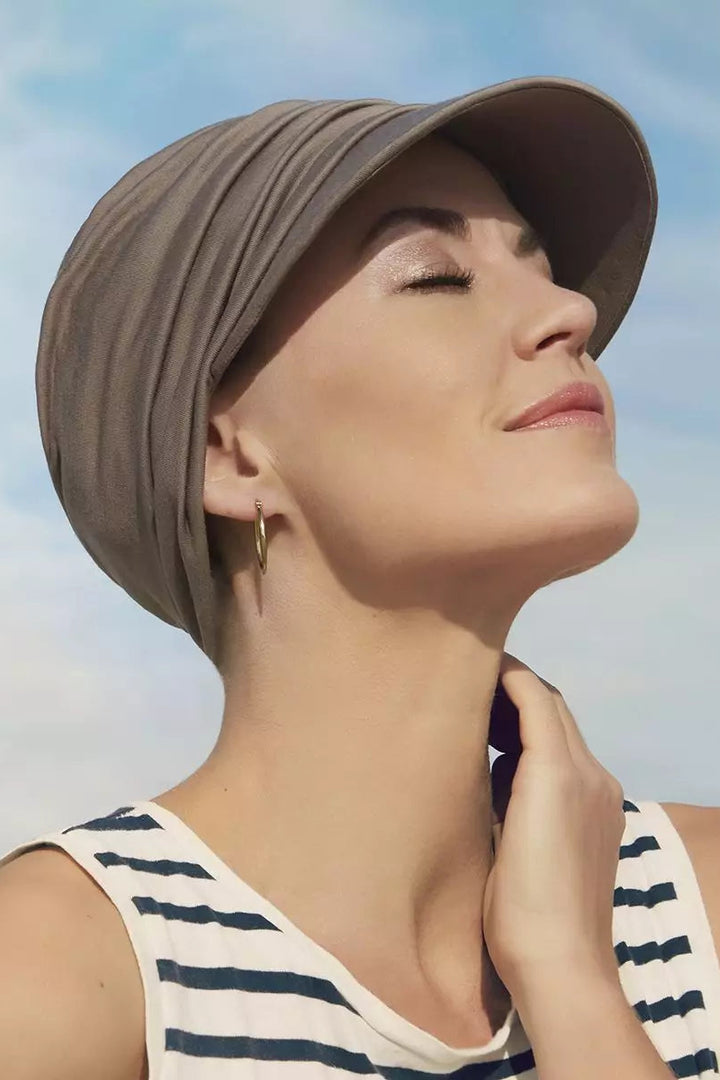 A woman wearing a taupe pleated hair loss sun turban/hat and a navy and white striped tank top, looking upward against a light blue sky.