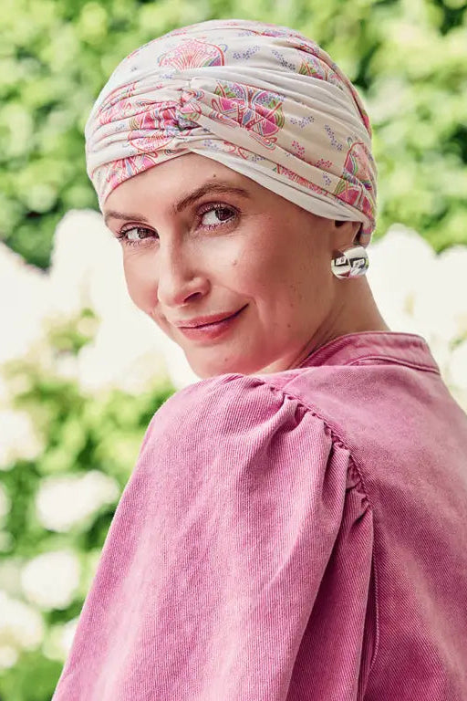 A woman outdoors wearing a pink and beige floral patterned headwrap turban and a dusty pink velvet puff-sleeve top, looking back over her shoulder.
